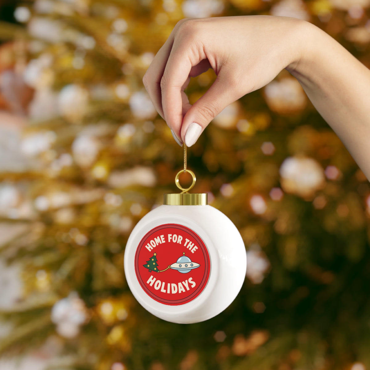 A hand holding a white spherical Christmas ornament hanging from a tree branch, featuring a red circular graphic with the text 'HOME FOR THE HOLIDAYS' and a cartoon UFO pulling a Christmas tree.