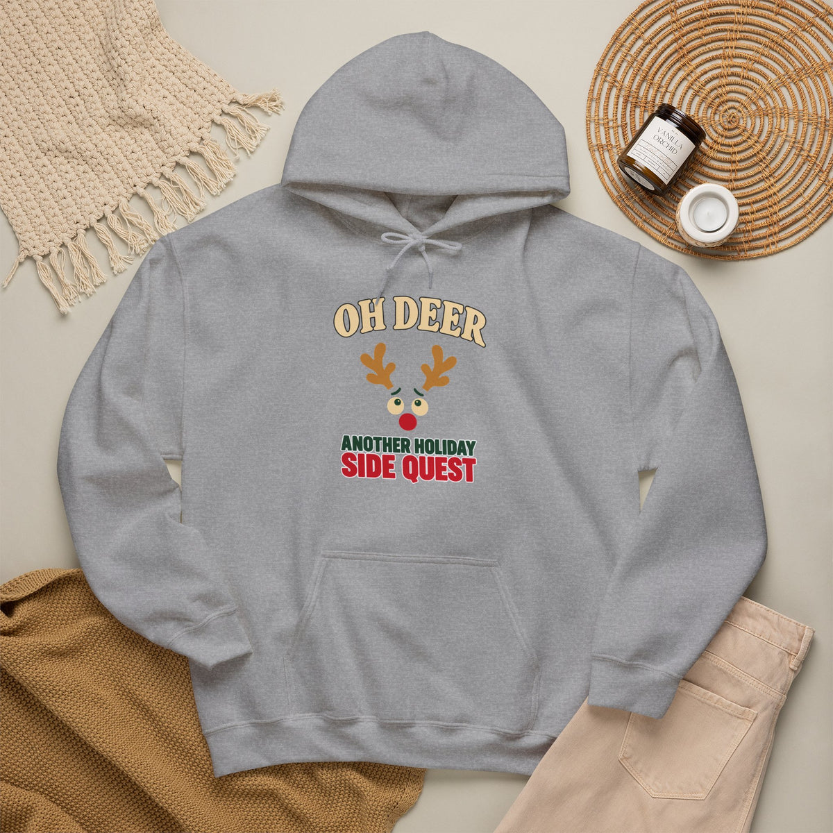 A light gray pullover hoodie laid flat with a front kangaroo pocket and a printed chest graphic reading "OH DEER" above and below ' ANOTHER HOLIDAY SIDE QUEST The hoodie is styled in a flat lay with a woven blanket, tan knit fabric, a small jar, a round tray, and a small cup on a light neutral background.