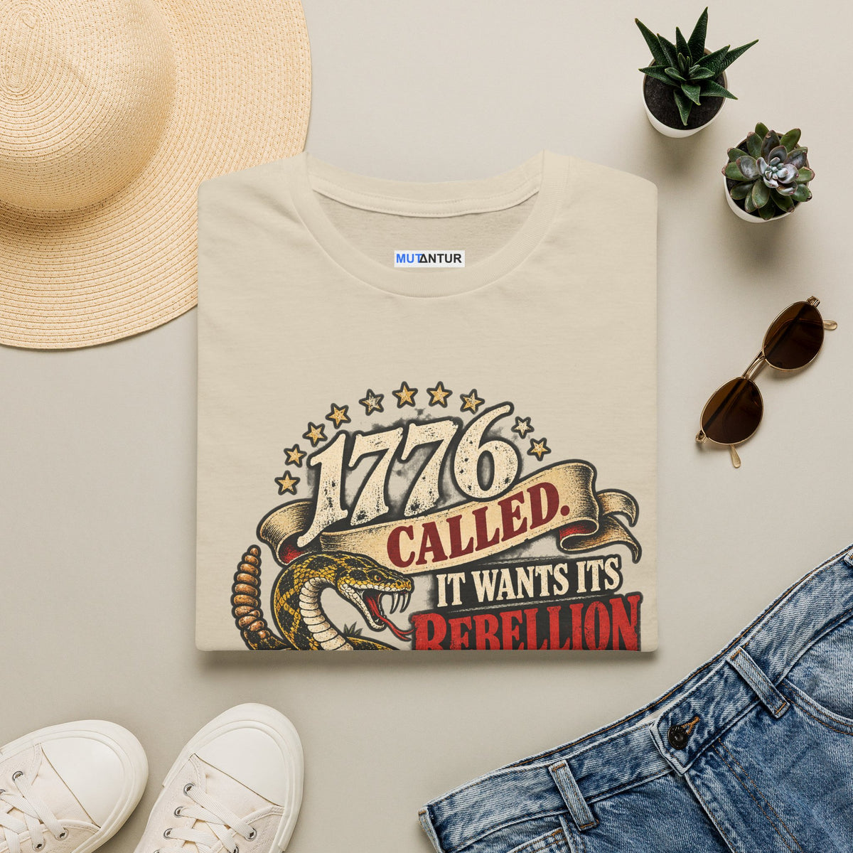 A folded cream-colored t-shirt with a vintage-style graphic reading '1776 CALLED. IT WANTS ITS REBELLION' with a snake illustration, styled in a flat lay with a straw hat, white sneakers, blue jeans, sunglasses, and small potted succulents on a light gray background.