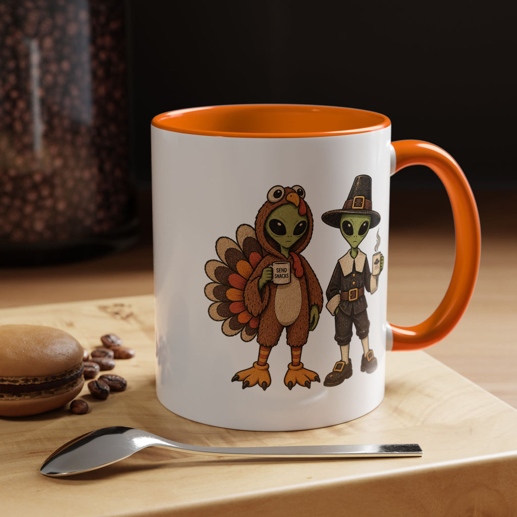 A white ceramic mug with an orange handle and interior, featuring a cartoon illustration of a turkey and a pilgrim on the side. The mug is set on a wooden surface next to a spoon and a macaron, with a blurred background.