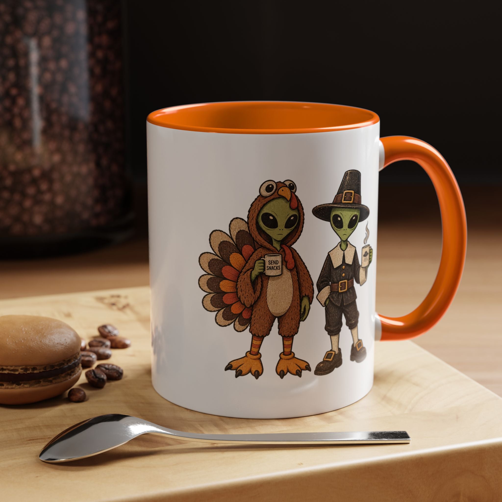 A white ceramic mug with an orange handle and interior, featuring a cartoon illustration of a turkey and a pilgrim on the side. The mug is set on a wooden surface next to a spoon and a macaron, with a blurred background.