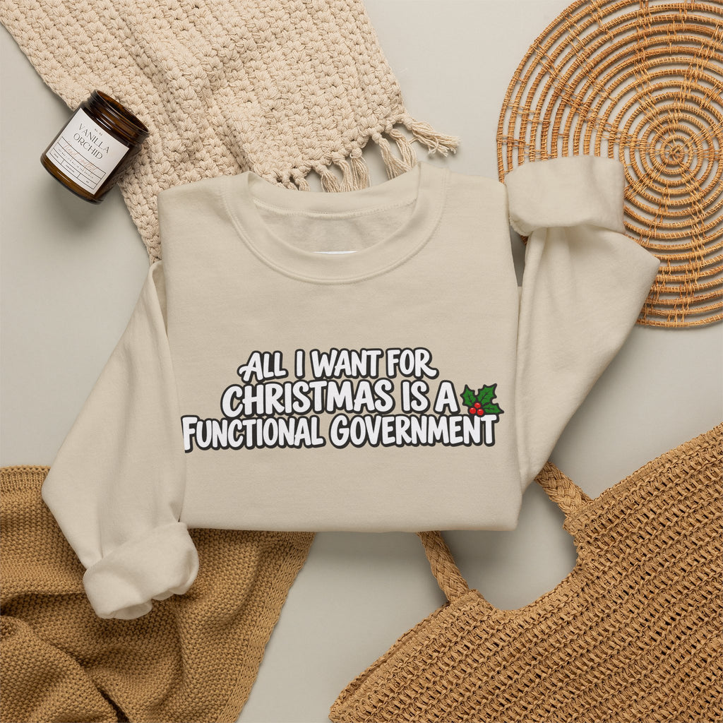 A folded cream sweatshirt with the text 'ALL I WANT FOR CHRISTMAS IS A FUNCTIONAL GOVERNMENT' and a small holly graphic, styled in a flat lay with a woven blanket, a small candle jar, a wicker tray, and a tan knit bag on a light surface.