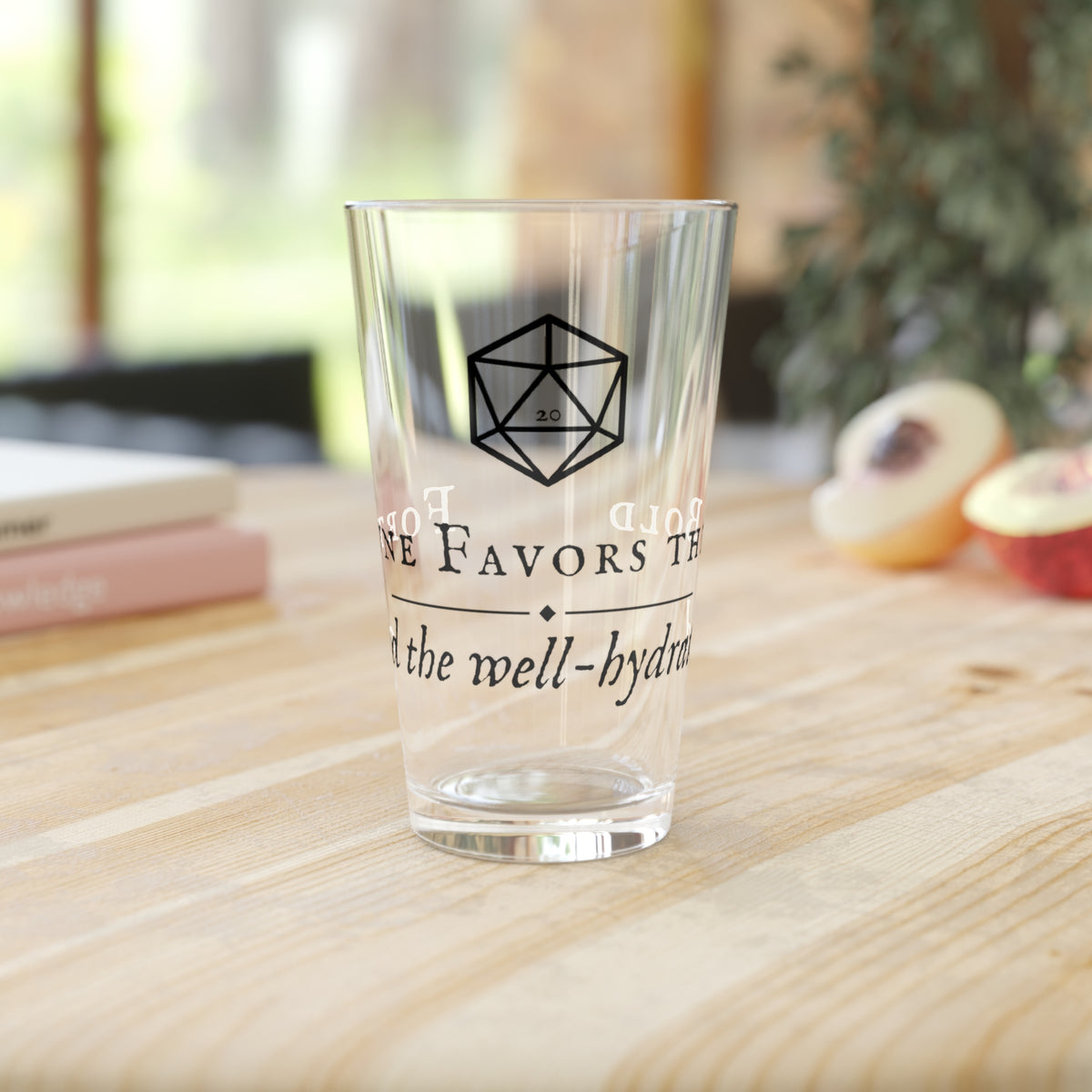 A clear drinking glass with black printed text and a geometric logo, standing on a light wood tabletop in a softly blurred indoor setting.