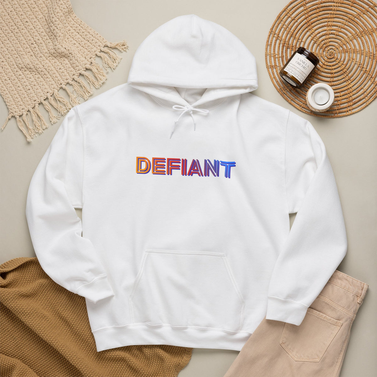 Defiant rainbow text logo graphic hoodie in white, flat lay styled with a knit blanket, coaster, and jar