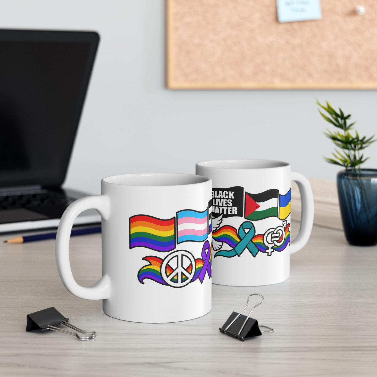 Two white ceramic mugs with colorful pride and social justice graphics, displayed on a light wood desk with a laptop and plant in the background.