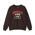 A dark brown crewneck sweatshirt featuring a retro VHS graphic on the chest that reads 'CHRISTMAS ON VHS' with 'BE KIND REWIND' underneath, displayed on a clean white background.