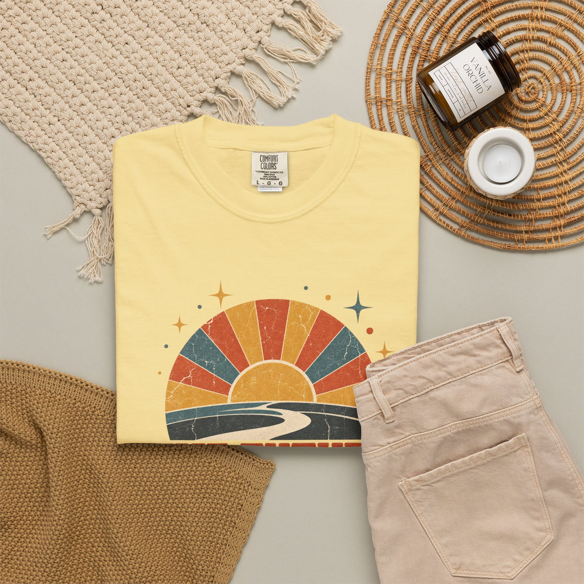 A folded pale yellow T-shirt with a retro sunset graphic in orange, red, blue, and yellow, styled in a flat lay with beige pants, a woven placemat, a candle jar, and a small cup on a light gray surface.