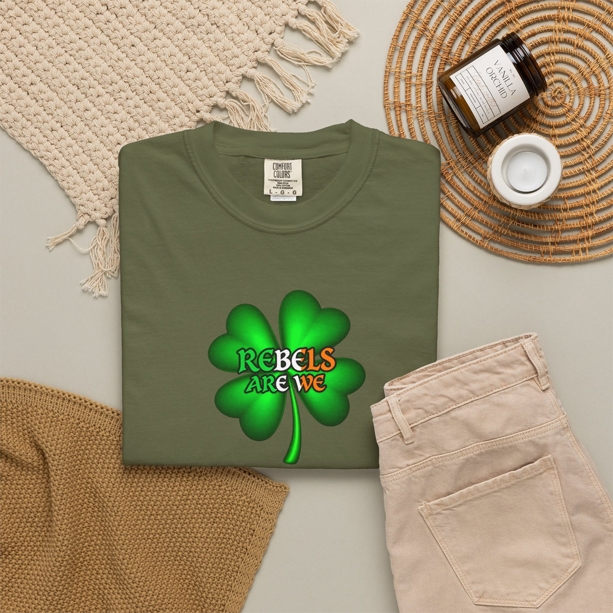 A folded olive green t-shirt with a green shamrock graphic and the text 'REBELS ARE WE' in red, yellow, and green, styled in a flat lay with beige pants, a woven blanket, a candle jar, and a small cup on a light gray background.