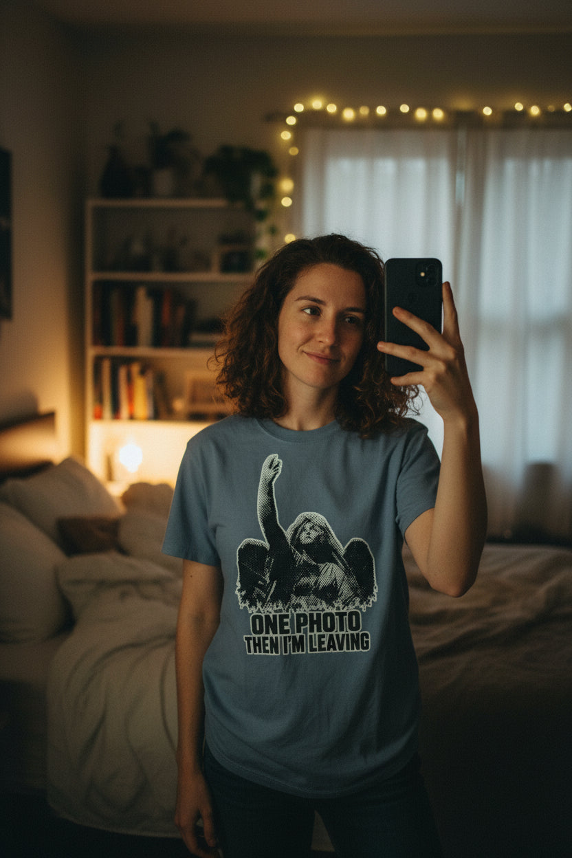 A slate blue crewneck t-shirt with a black and white graphic of a winged angel raising its arm, above the text 'ONE PHOTO THEN I'M LEAVING'.