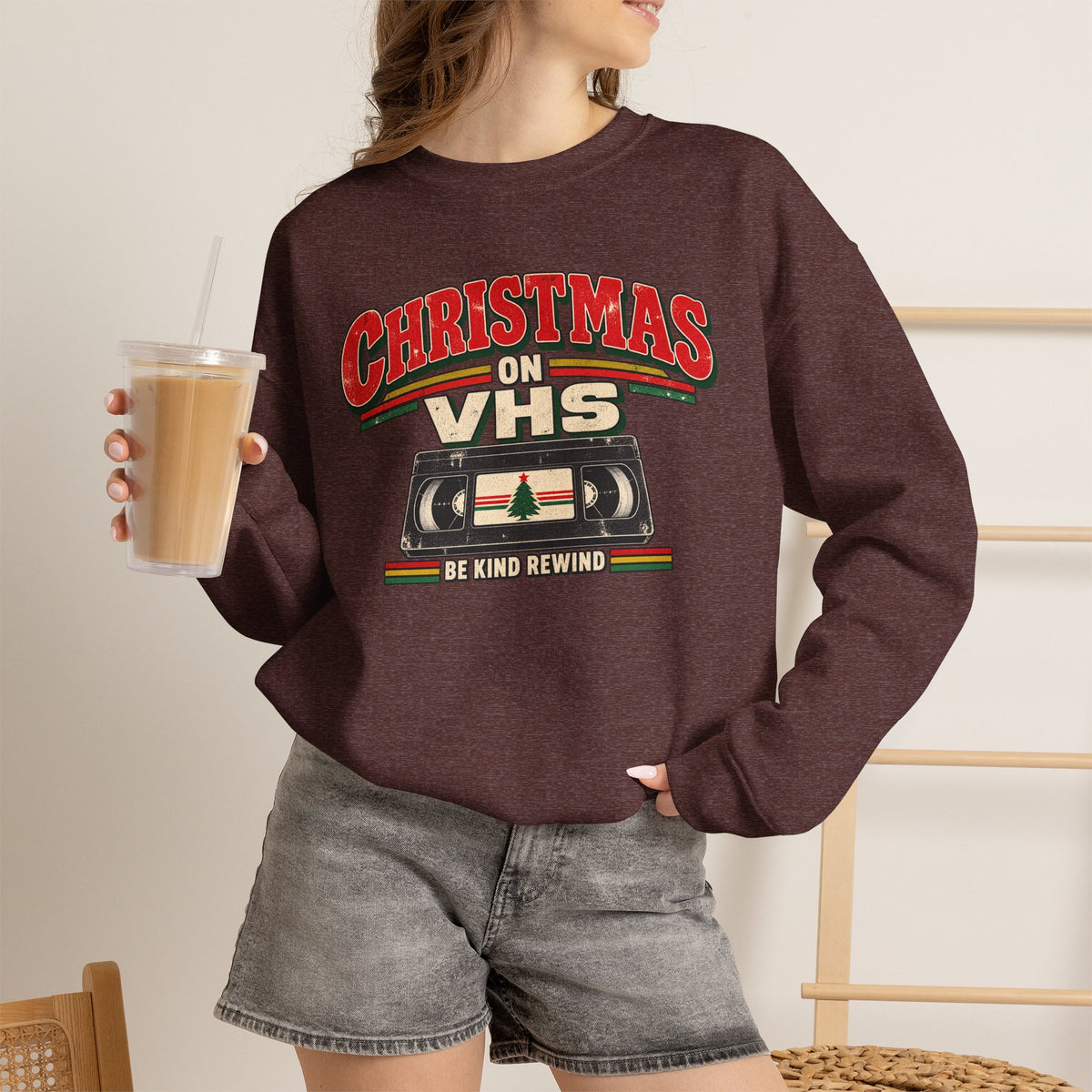 A woman wearing a dark heather brown crewneck sweatshirt with a retro 'Christmas on VHS' graphic, holding a iced coffee in a home setting.