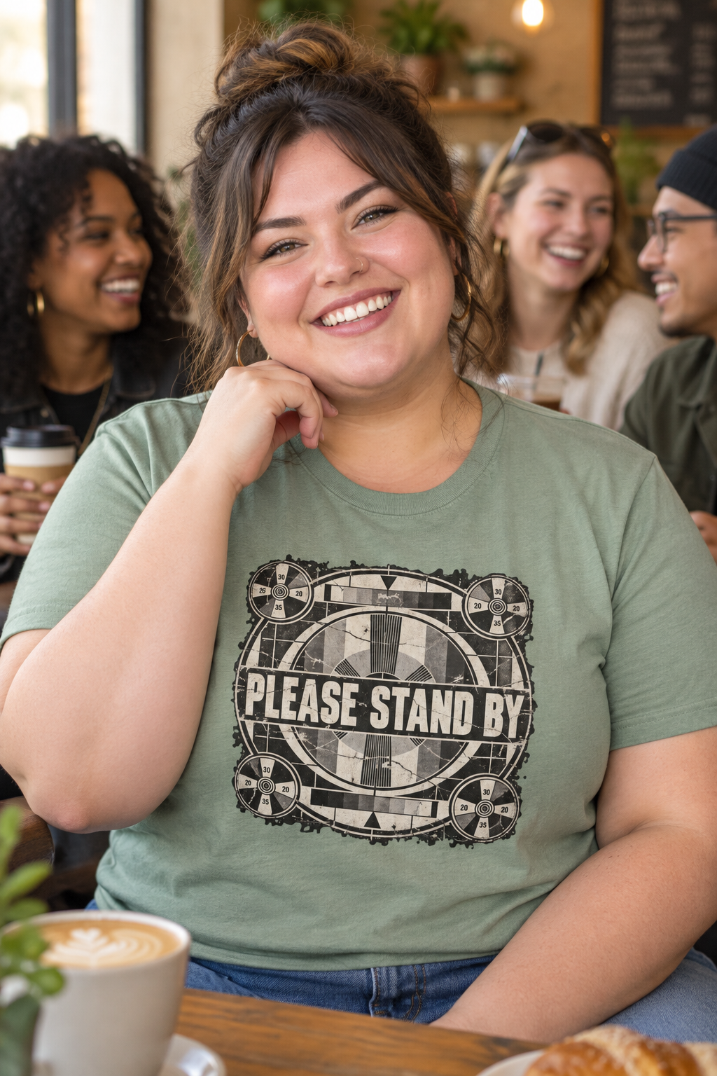 A woman smiling in a cafe setting, wearing an olive green graphic t-shirt with a black and white 'PLEASE STAND BY' design. Other people and coffee cups are visible in the background.