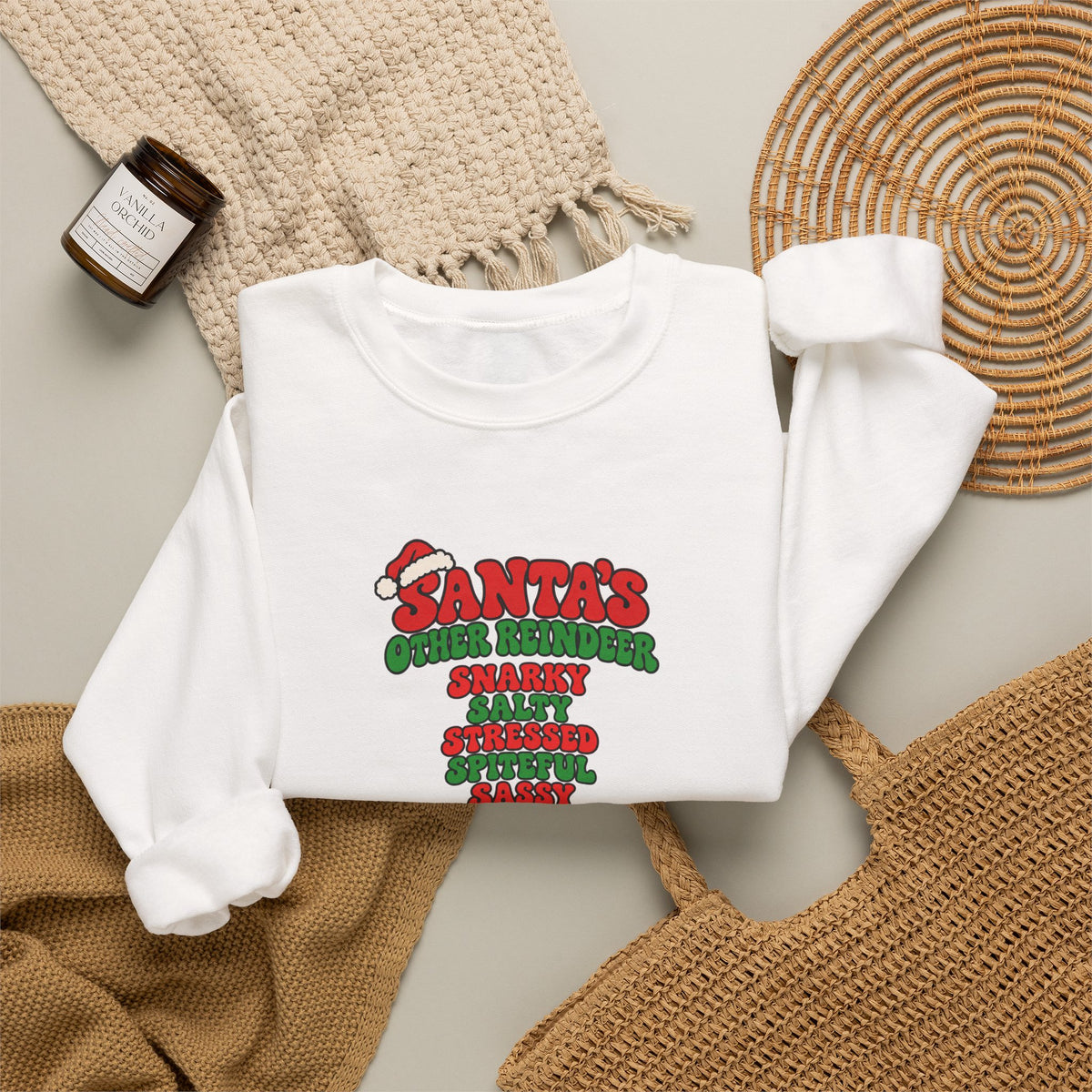 A folded white crewneck sweatshirt featuring a red and green graphic print on the front that reads 'SANTA'S OTHER REiNDEER' followed by a list of humorous qualities in red and green text, shown against a beige background