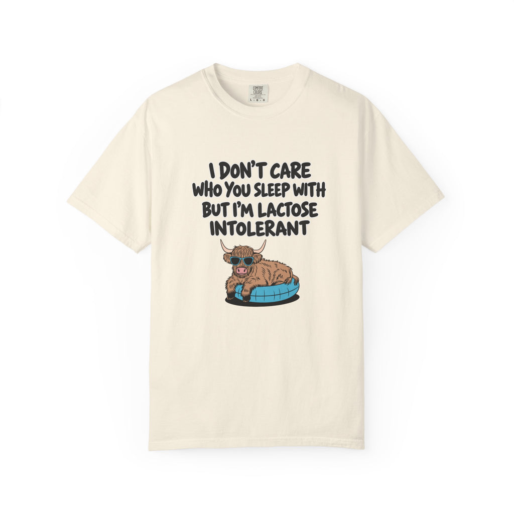 An off-white short-sleeve t-shirt with a graphic print of a cartoon Highland cow sitting on a blue tire. The text above the cow reads, 'I DON'T CARE WHO YOU SLEEP WITH BUT I'M LACTOSE INTOLERANT' in a black hand-drawn font.
