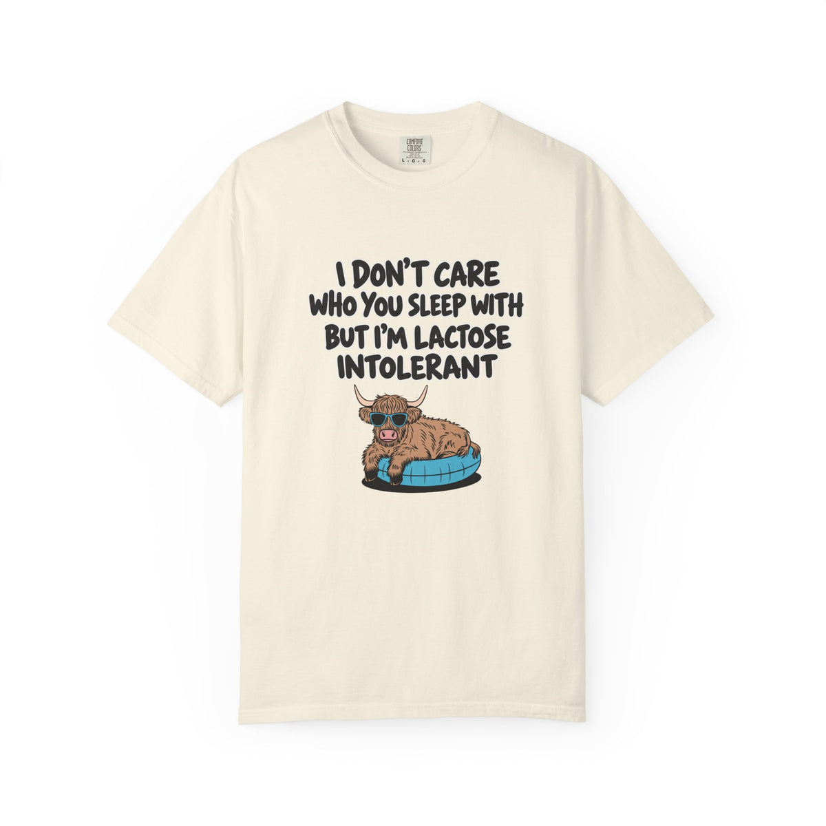 An off-white short-sleeve t-shirt with a graphic print of a cartoon Highland cow sitting on a blue tire. The text above the cow reads, 'I DON'T CARE WHO YOU SLEEP WITH BUT I'M LACTOSE INTOLERANT' in a black hand-drawn font.