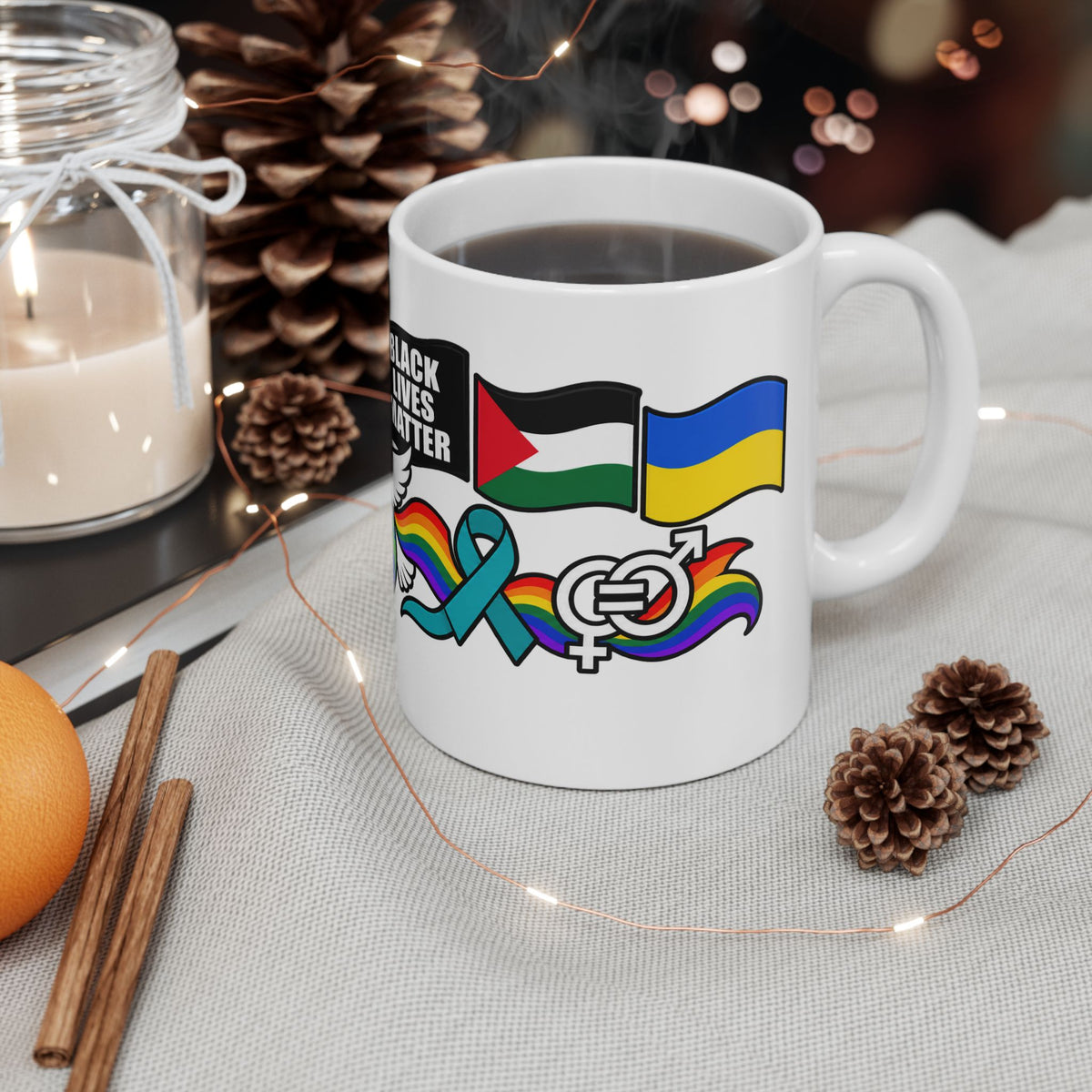 A white ceramic coffee mug with a black and white graphic reading 'BLACK LIVES MATTER' alongside Palestinian, Ukrainian, and rainbow pride ribbons and flags, shown on a light fabric surface with a candle, pinecones, cinnamon sticks, and fairy lights in the background.