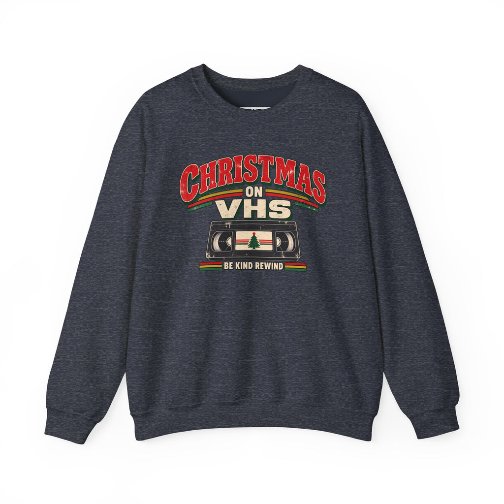 A dark heather gray crewneck sweatshirt featuring a retro Christmas graphic on the chest that reads 'CHRISTMAS ON VHS' in red and cream lettering above a cassette tape illustration, with 'BE KIND REWIND' underneath.