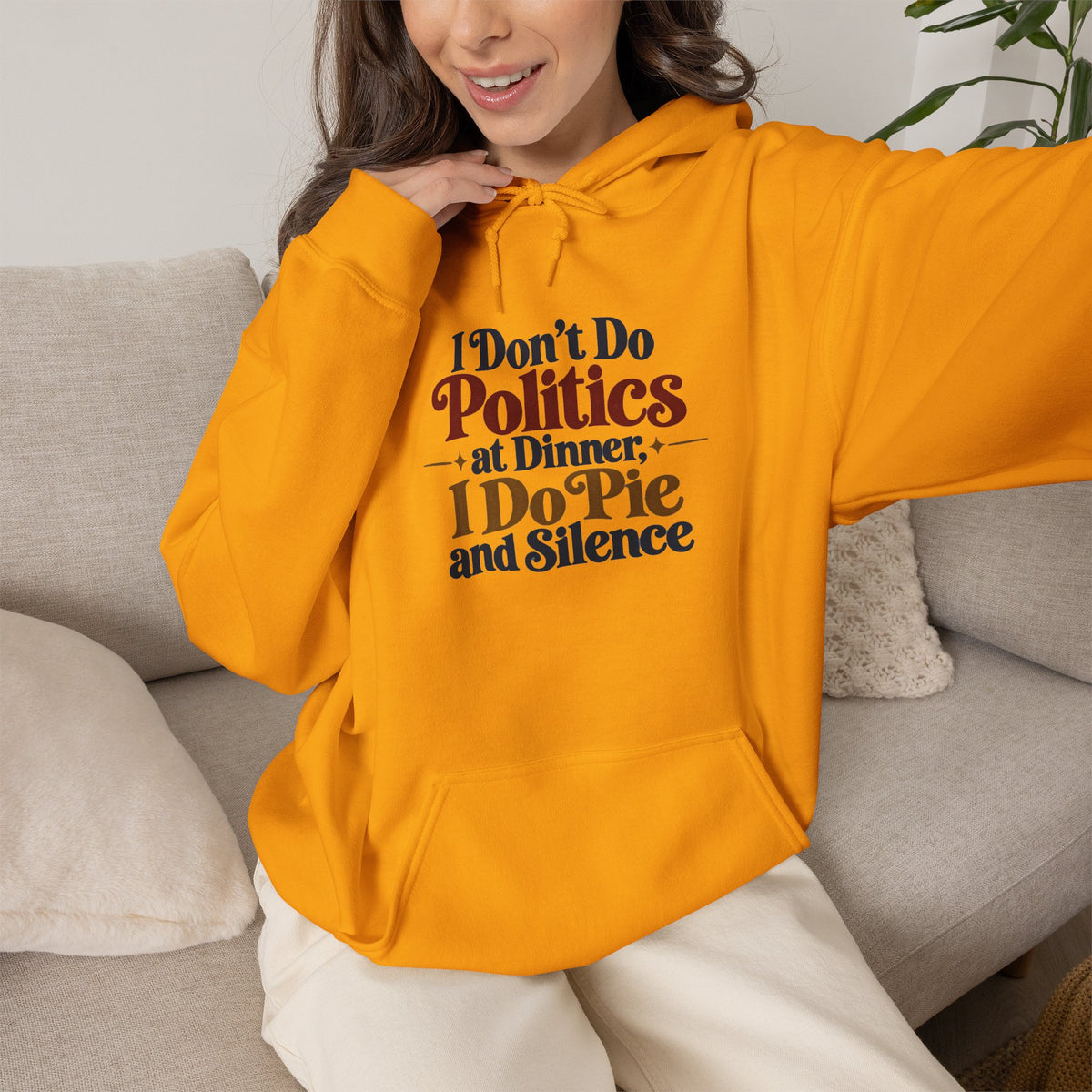 A woman wearing a bright yellow hoodie with the text 'I Don't Do Politics at Dinner, I Do Pie and Silence' in dark red and navy lettering, sitting on a light gray couch.
