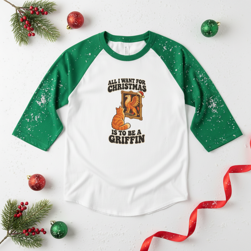 A raglan-style baseball T-shirt with green sleeves and a white body, decorated with a Christmas-themed graphic that reads 'ALL I WANT FOR CHRISTMAS IS TO BE A GRIFFIN' with an orange cat and a golden bird illustration. The shirt is styled in a flat lay with pine branches, red ornaments, and a red ribbon on a white background.