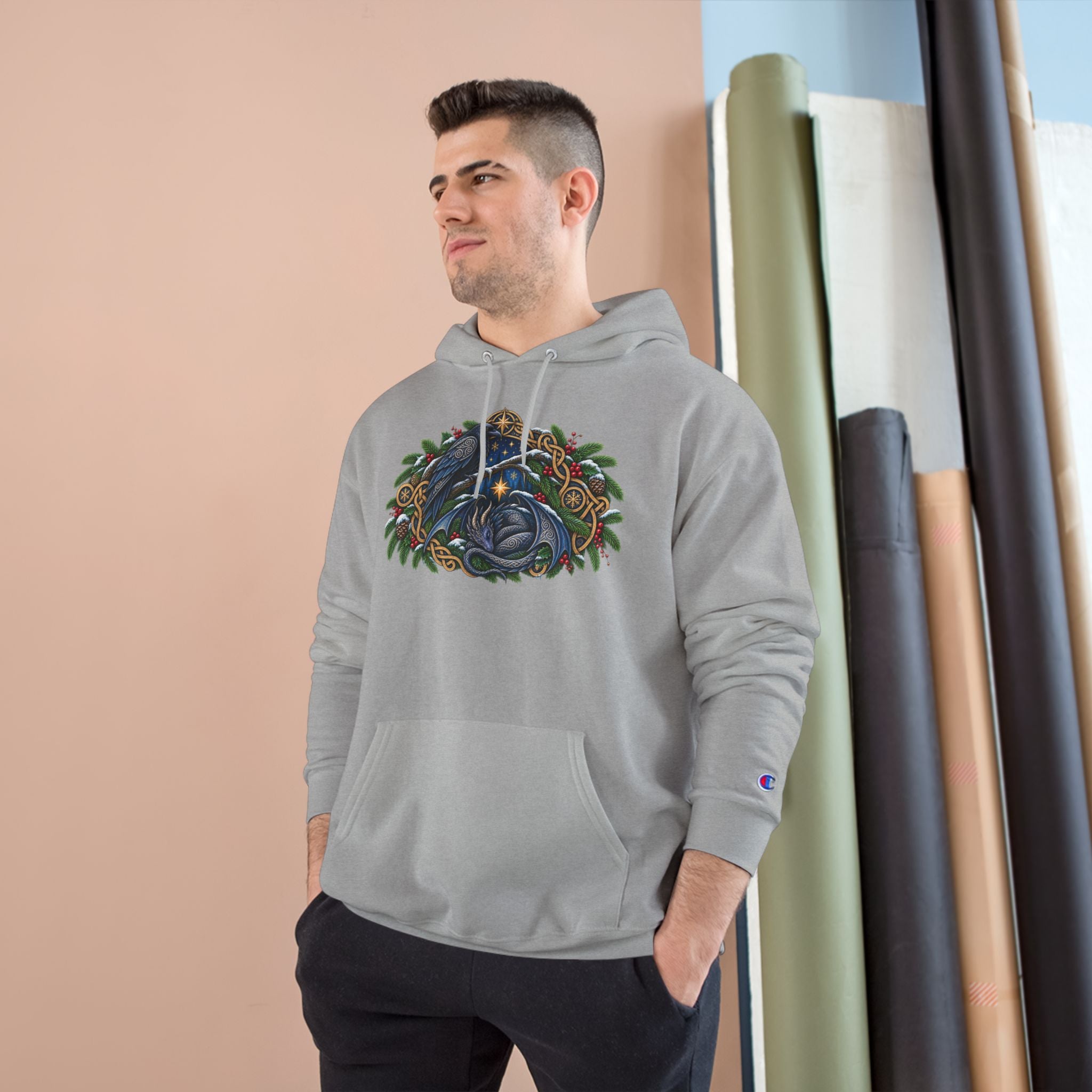 a man wearing a light gray pullover hoodie featuring a detailed graphic of a blue raven and a dragon surrounded by green pine boughs  A small blue Champion 'C' logo is visible on the left sleeve cuff. 