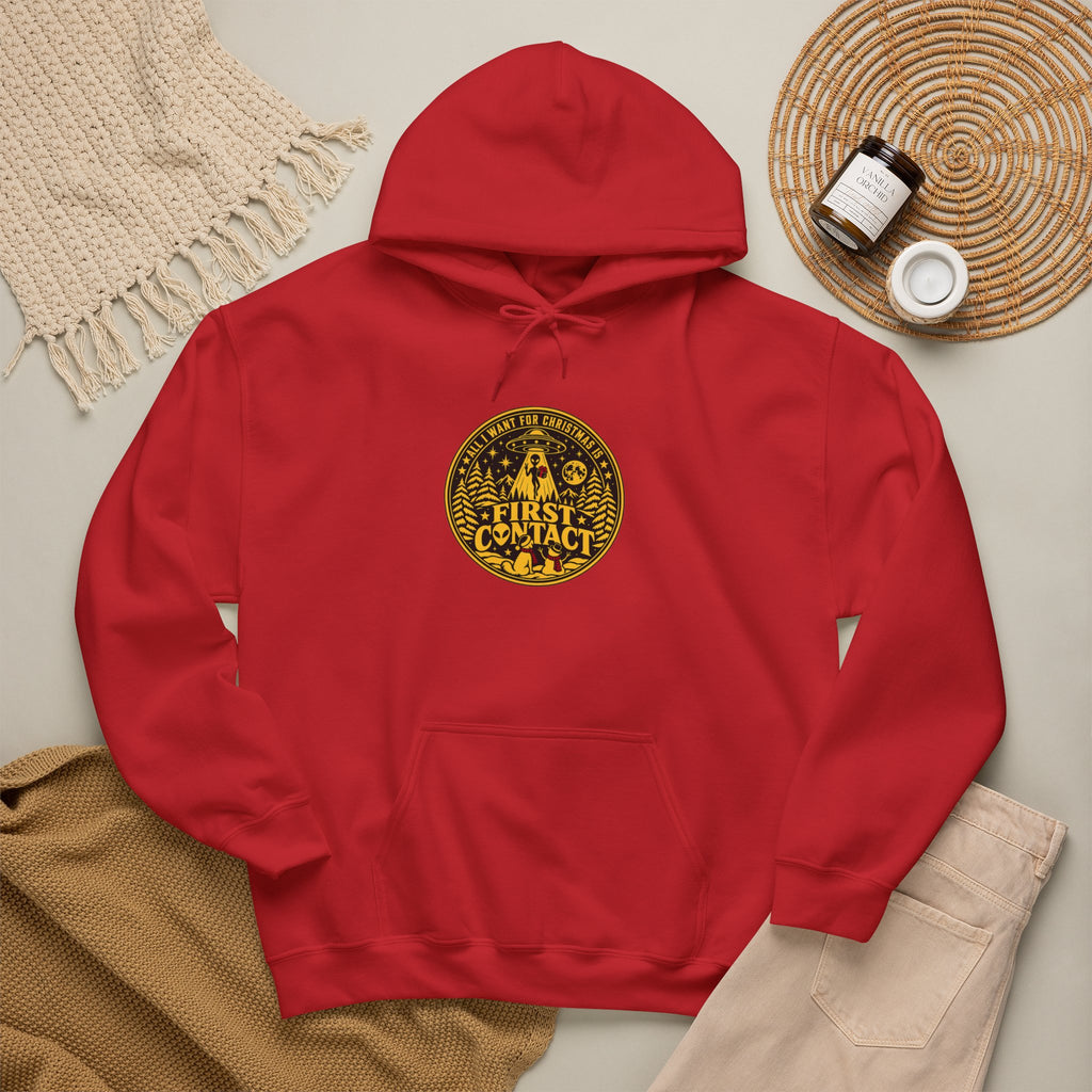 A red pullover hoodie laid flat on a light beige surface, featuring a circular yellow and black graphic on the chest that reads All I want for Christmas is  'FIRST CONTACT' with a dog and a reindeer looking up at an alien rising into a UFO at Christmas . The hoodie is styled with a woven blanket, tan fabric, a small jar, and a small cup in a flat-lay arrangement.