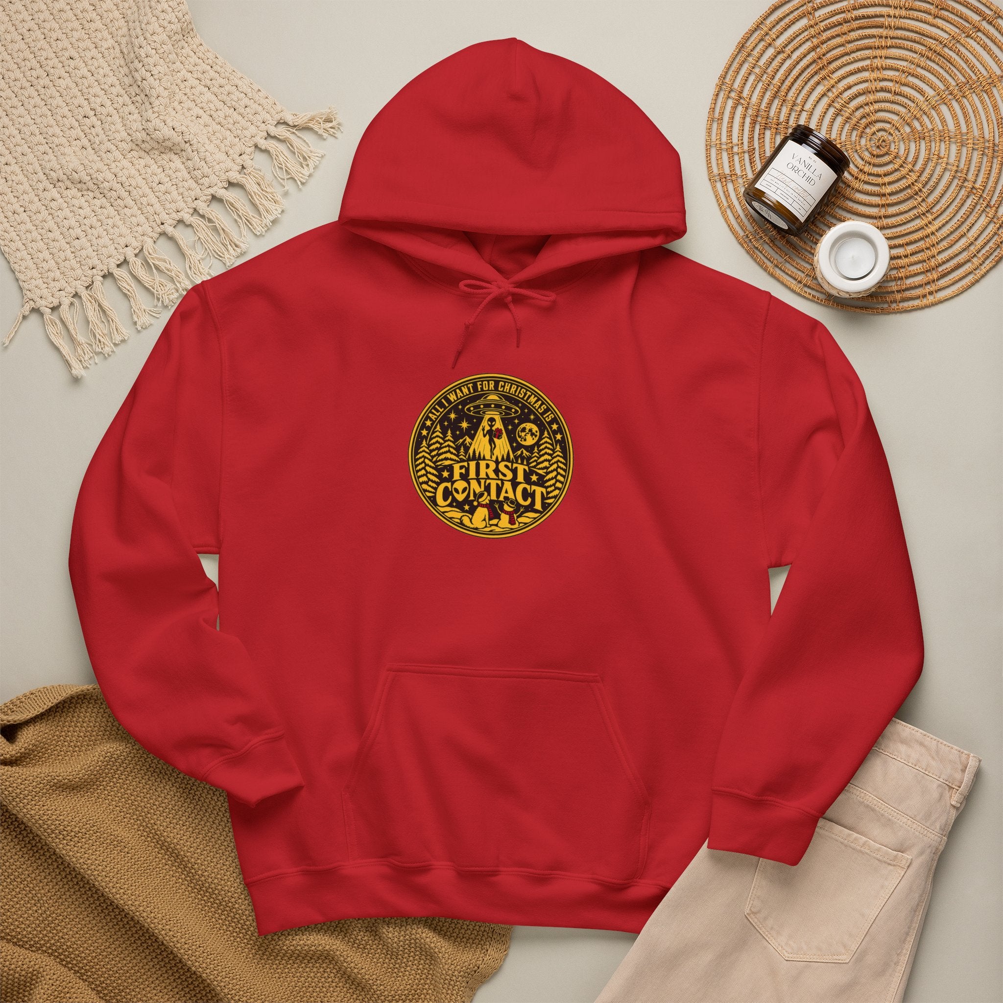 A red pullover hoodie laid flat on a light beige surface, featuring a circular yellow and black graphic on the chest that reads All I want for Christmas is  'FIRST CONTACT' with a dog and a reindeer looking up at an alien rising into a UFO at Christmas . The hoodie is styled with a woven blanket, tan fabric, a small jar, and a small cup in a flat-lay arrangement.