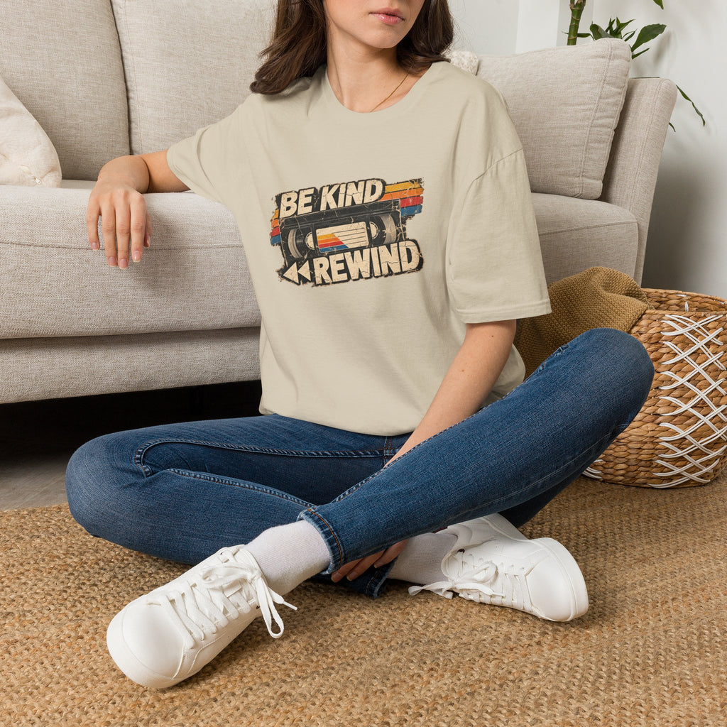 A woman sitting cross-legged on a woven rug in front of a light beige sofa, wearing a beige graphic T-shirt with a retro cassette tape design and the text 'BE KIND REWIND', blue jeans, and white sneakers.