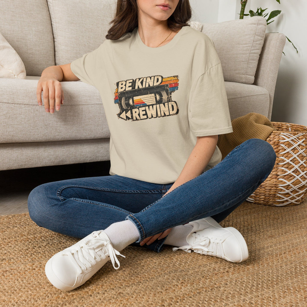 A woman sitting cross-legged on a woven rug in front of a light beige sofa, wearing a beige graphic T-shirt with a retro cassette tape design and the text 'BE KIND REWIND', blue jeans, and white sneakers.