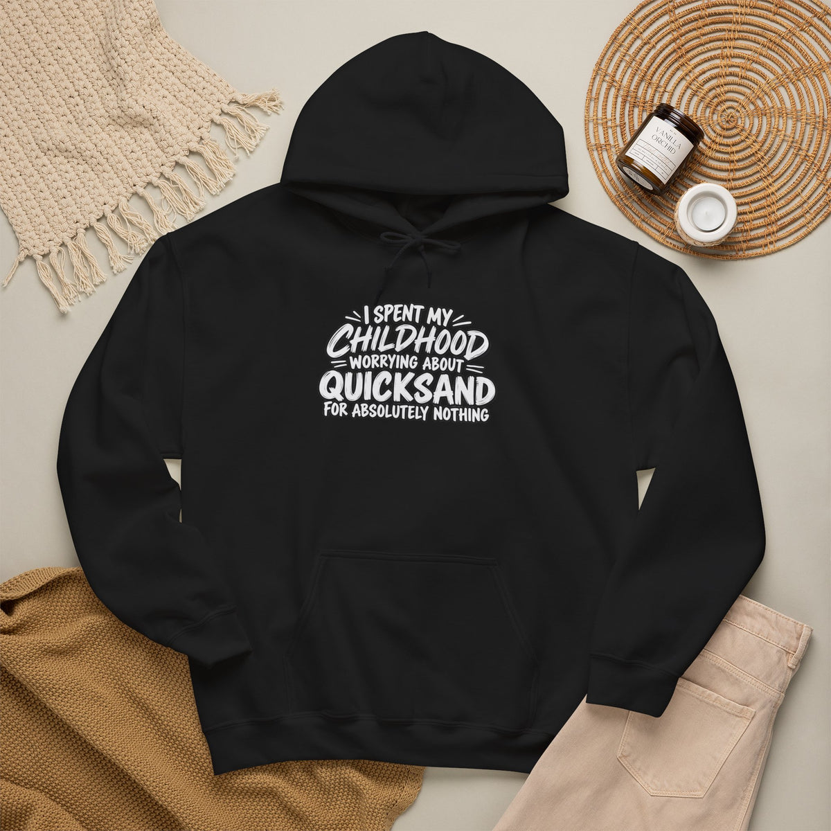 black hoodie with white text that reads: I SPEND MY CHILDHOOD WORRYING ABOUT QUICKSAND FOR ABSOLUTELY NOTHING. it is displayed with a beige knitted scarf and a candle