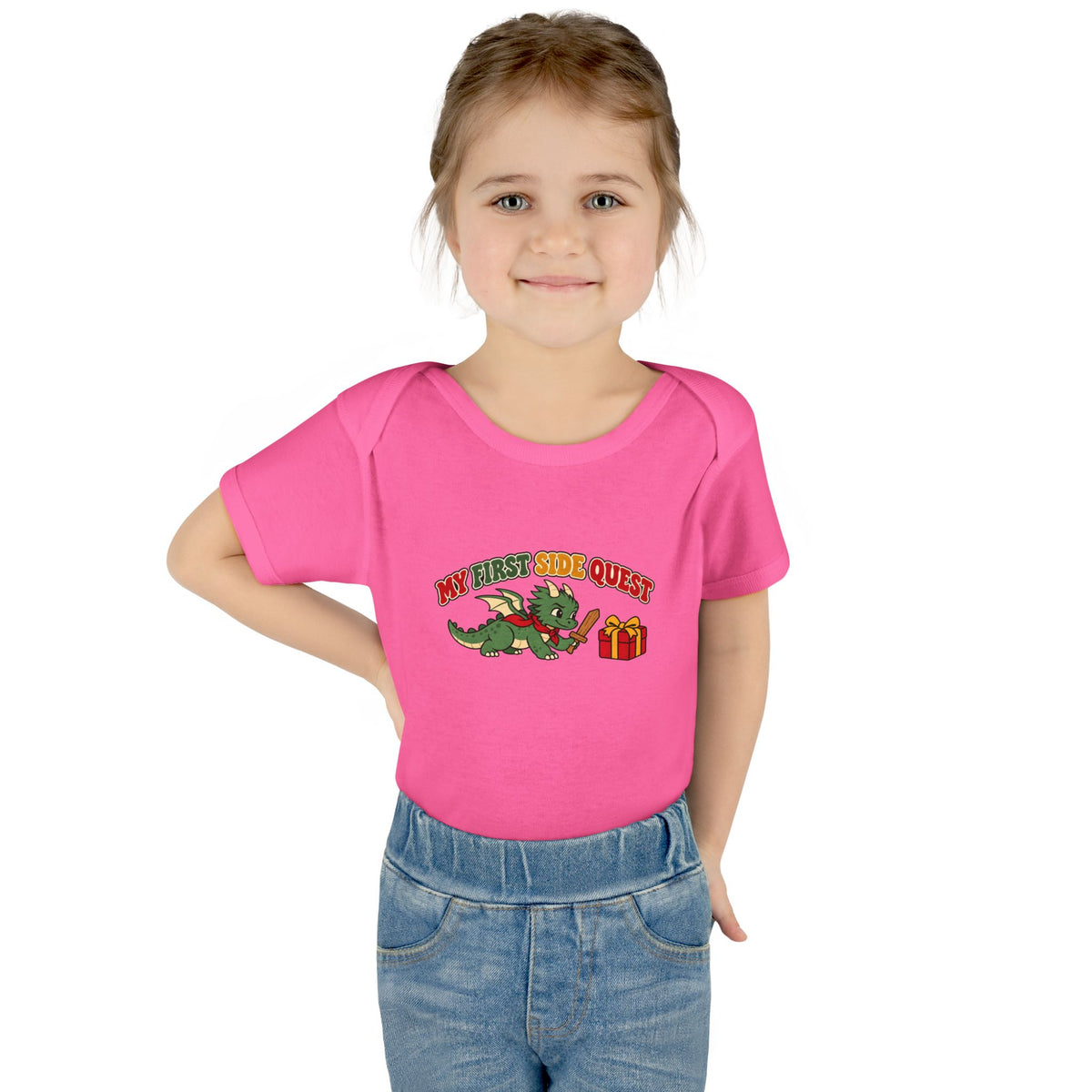 A young girl with light brown hair wearing a bright pink short-sleeve t-shirt with a cartoon graphic that reads 'MY FIRST SIDE QUEST' next to a green dragon and a red gift box. She is also wearing blue jeans, standing against a solid white background.