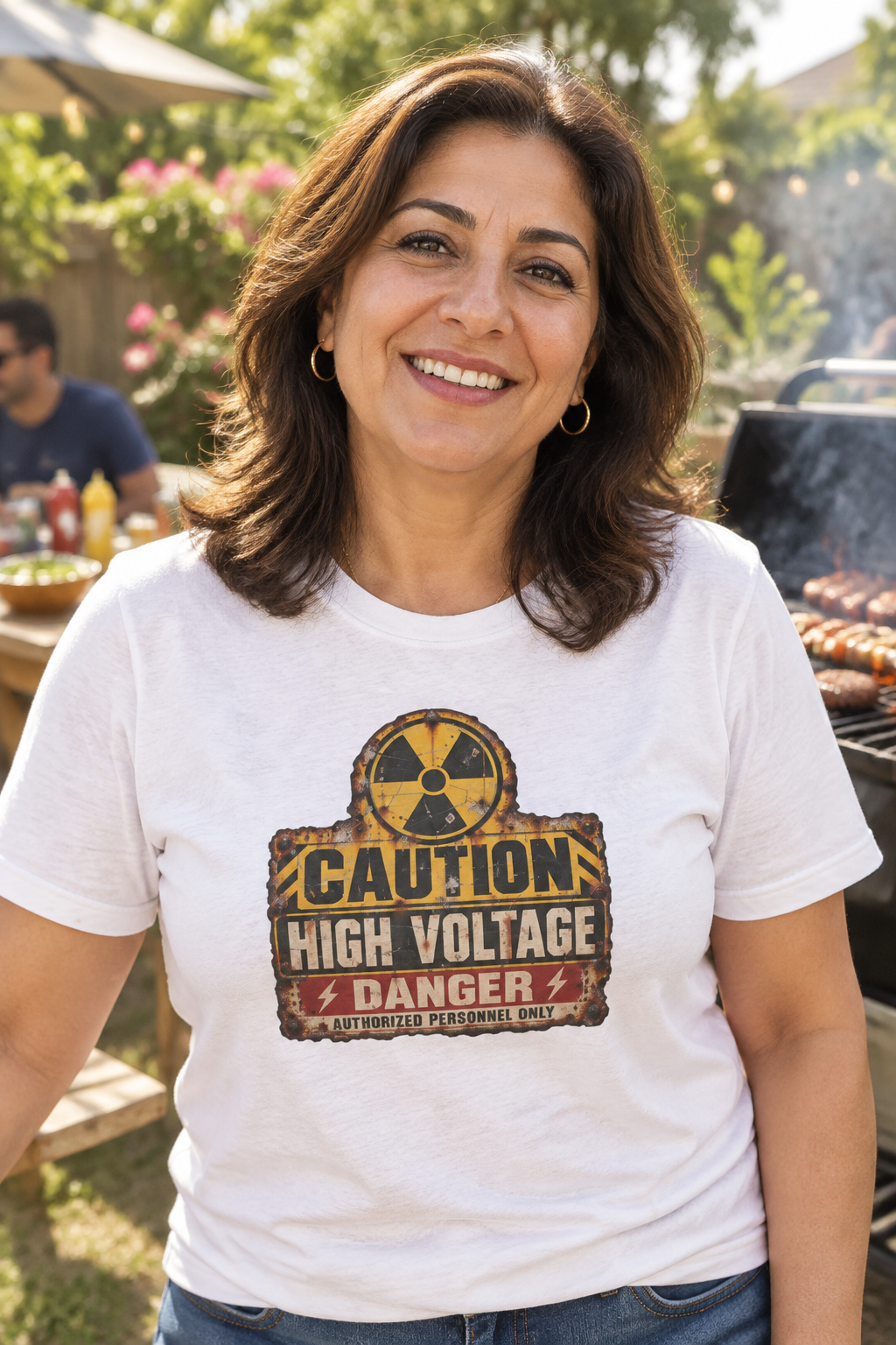 A smiling woman wearing a white graphic t-shirt with a 'CAUTION HIGH VOLTAGE DANGER' radiation symbol print, standing outdoors in a backyard grill setting.