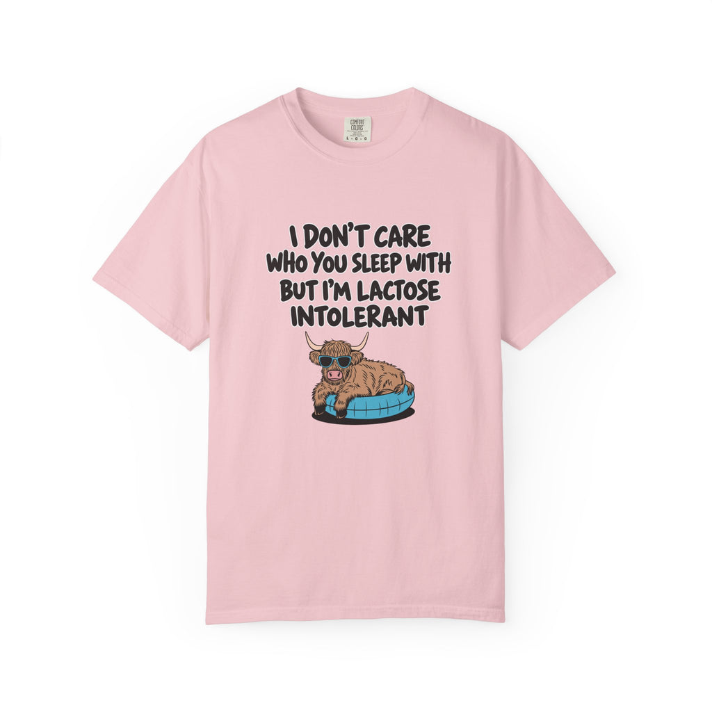 A light pink short-sleeve t-shirt with a humorous graphic on the front. The graphic features the text 'I DON'T CARE WHO YOU SLEEP WITH BUT I'M LACTOSE INTOLERANT' in black capital letters above an illustration of a brown highland cow lying on a blue and black tire.