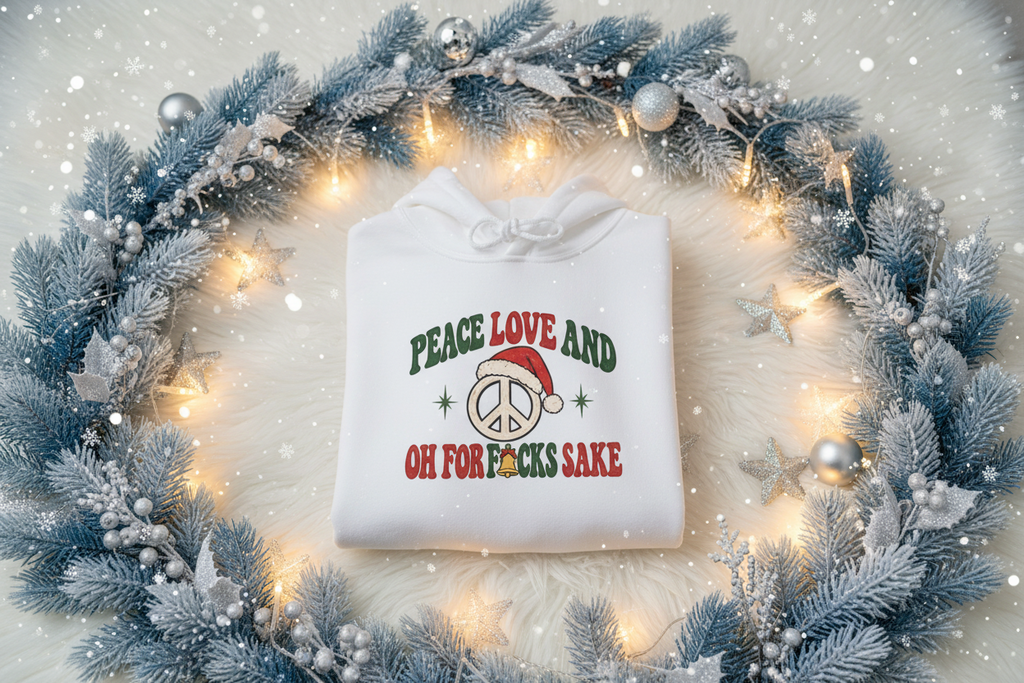 A folded white hoodie with a festive Christmas graphic reading 'PEACE LOVE AND OH FOR F*CKS SAKE' in green and red lettering with a Santa hat and peace sign, displayed on a white faux fur surface surrounded by a blue frosted wreath with warm string lights.