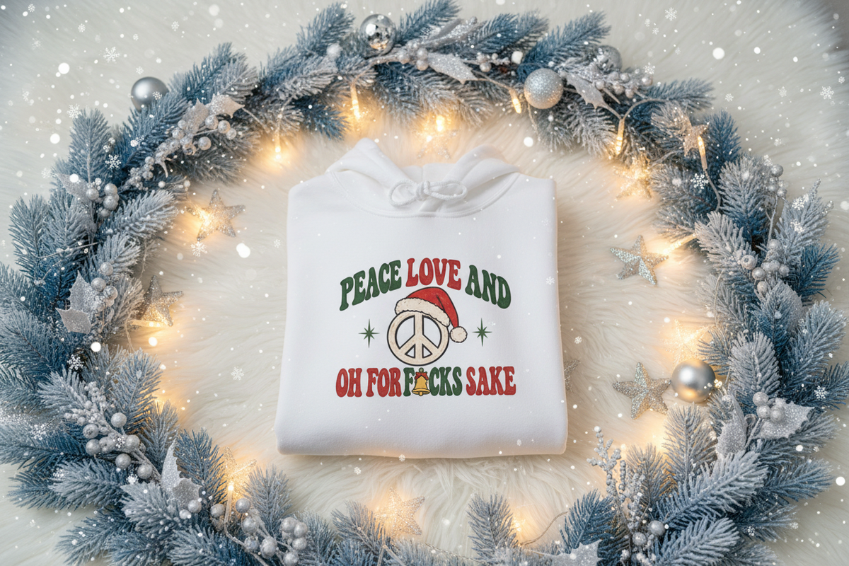 A folded white hoodie with a festive Christmas graphic reading 'PEACE LOVE AND OH FOR F*CKS SAKE' in green and red lettering with a Santa hat and peace sign, displayed on a white faux fur surface surrounded by a blue frosted wreath with warm string lights.