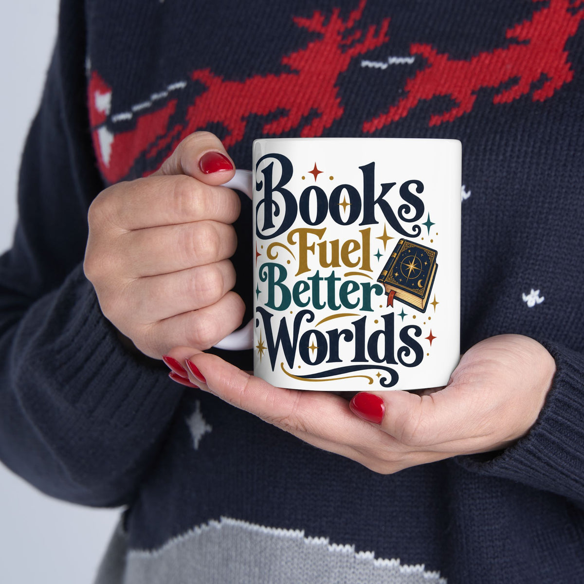 A person holding a white ceramic mug with the phrase 'Books Fuel Better Worlds' in stylized dark blue, gold, and teal lettering, wearing a navy knit sweater with a red and white pattern.