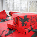Santa Dragon Magic Mayhem wrapping paper in red with black dragon silhouettes, wrapped gift box reading 'Magic Mayhem' on a table near a window, with paper roll and scissors