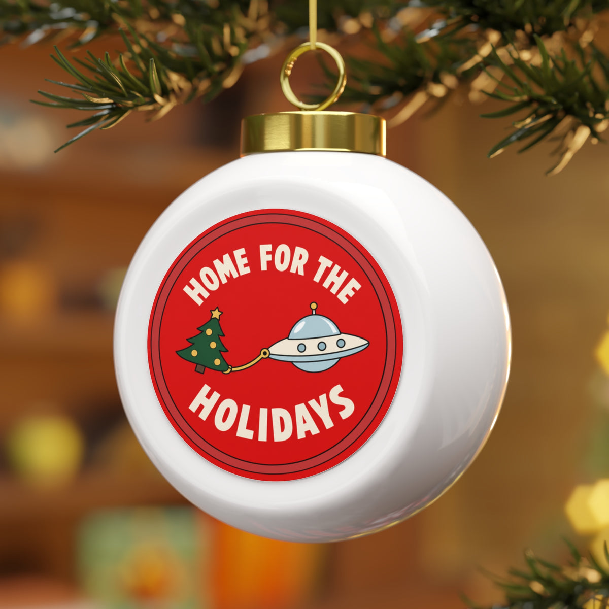 A white spherical Christmas ornament hanging from a tree branch, featuring a red circular graphic with the text 'HOME FOR THE HOLIDAYS' and a cartoon UFO pulling a Christmas tree.