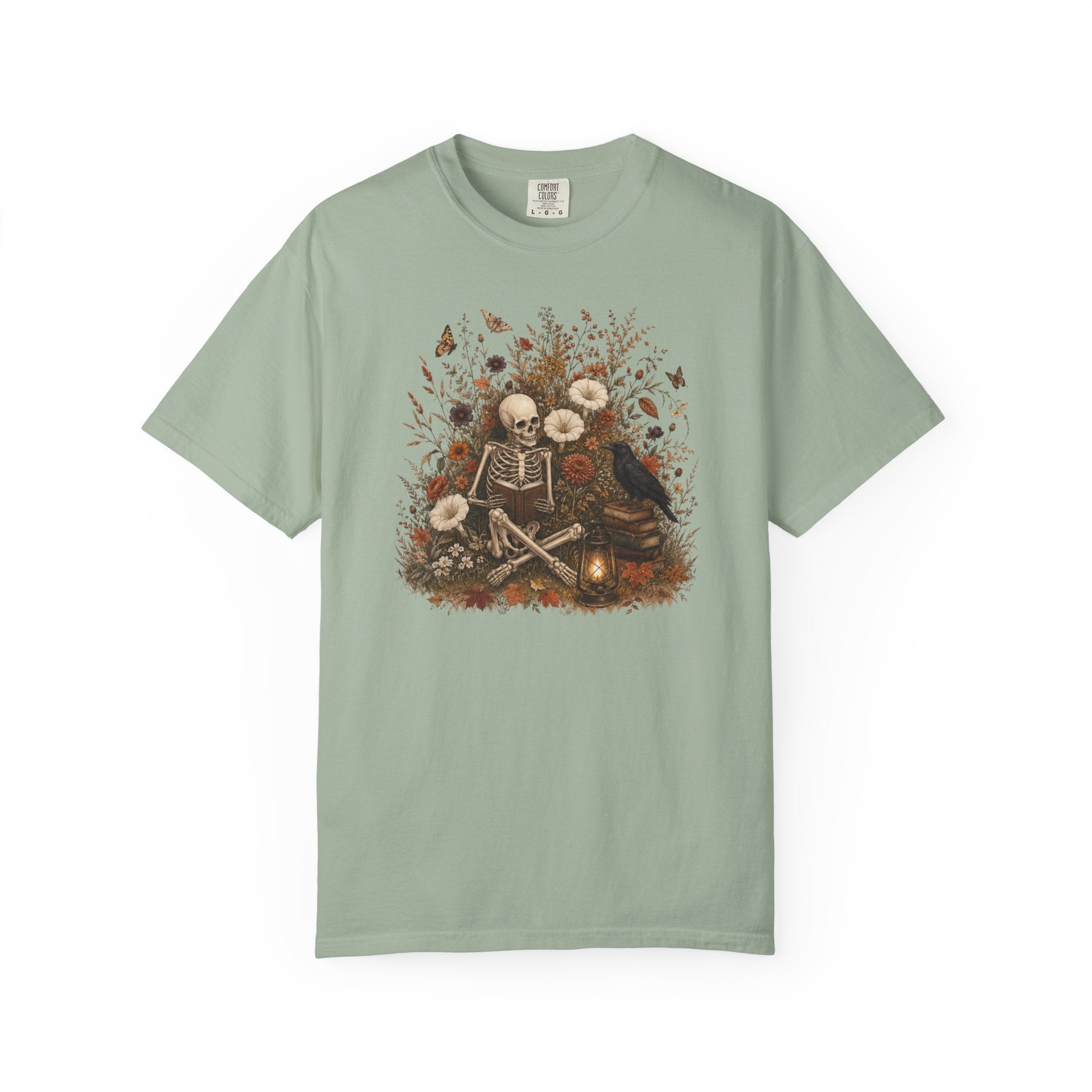 A sage green short-sleeve t-shirt with a detailed graphic print on the front showing a skeleton sitting by a campfire surrounded by flowers and a crow.