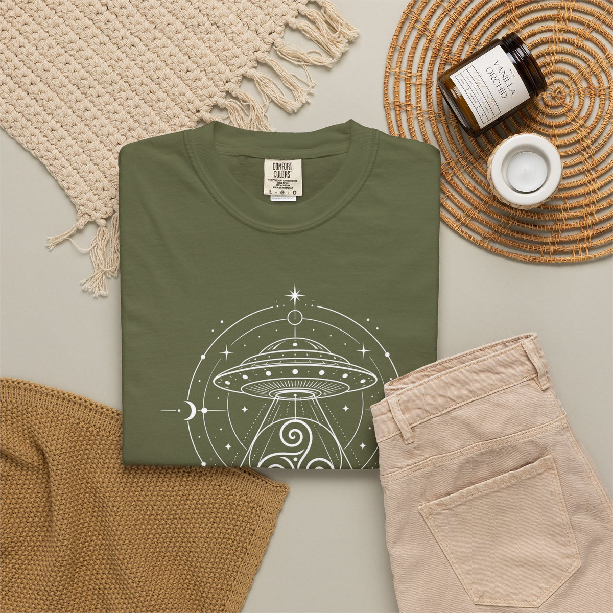 An olive green crewneck t-shirt with a white line-art graphic of a UFO and celestial pattern, styled in a flat lay with beige pants, a woven blanket, a candle, and a jar on a neutral background.