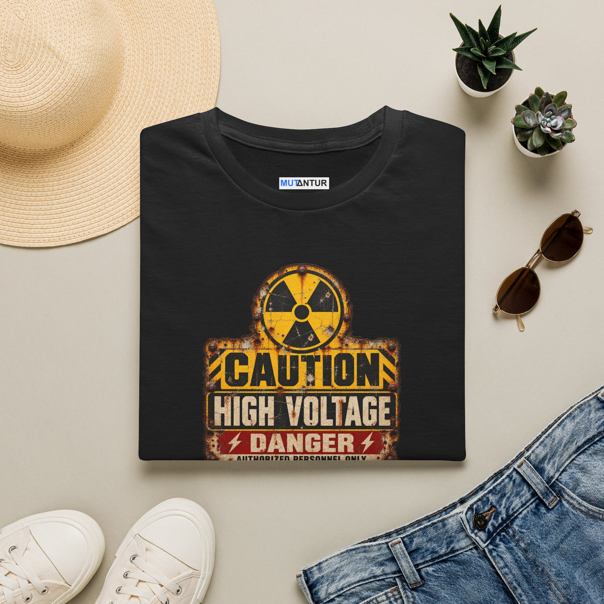 A folded black t-shirt with a distressed yellow, black, and red 'High Voltage' warning sign graphic on the front, displayed o with a sun hat sunglasses and other items of interest
