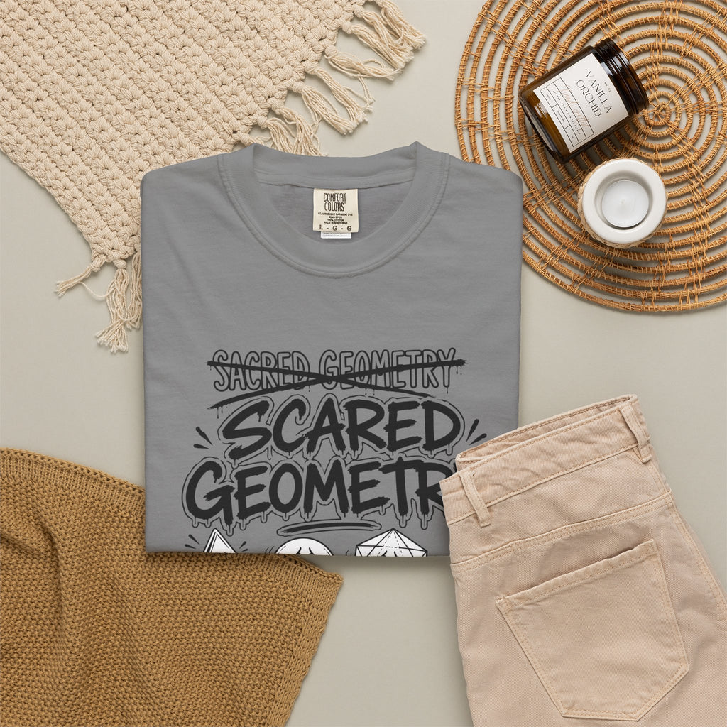 A folded gray 'Scared Geometry' graphic t-shirt with spooky-faced dice designs, styled flat on a knit blanket next to a pair of tan pants.