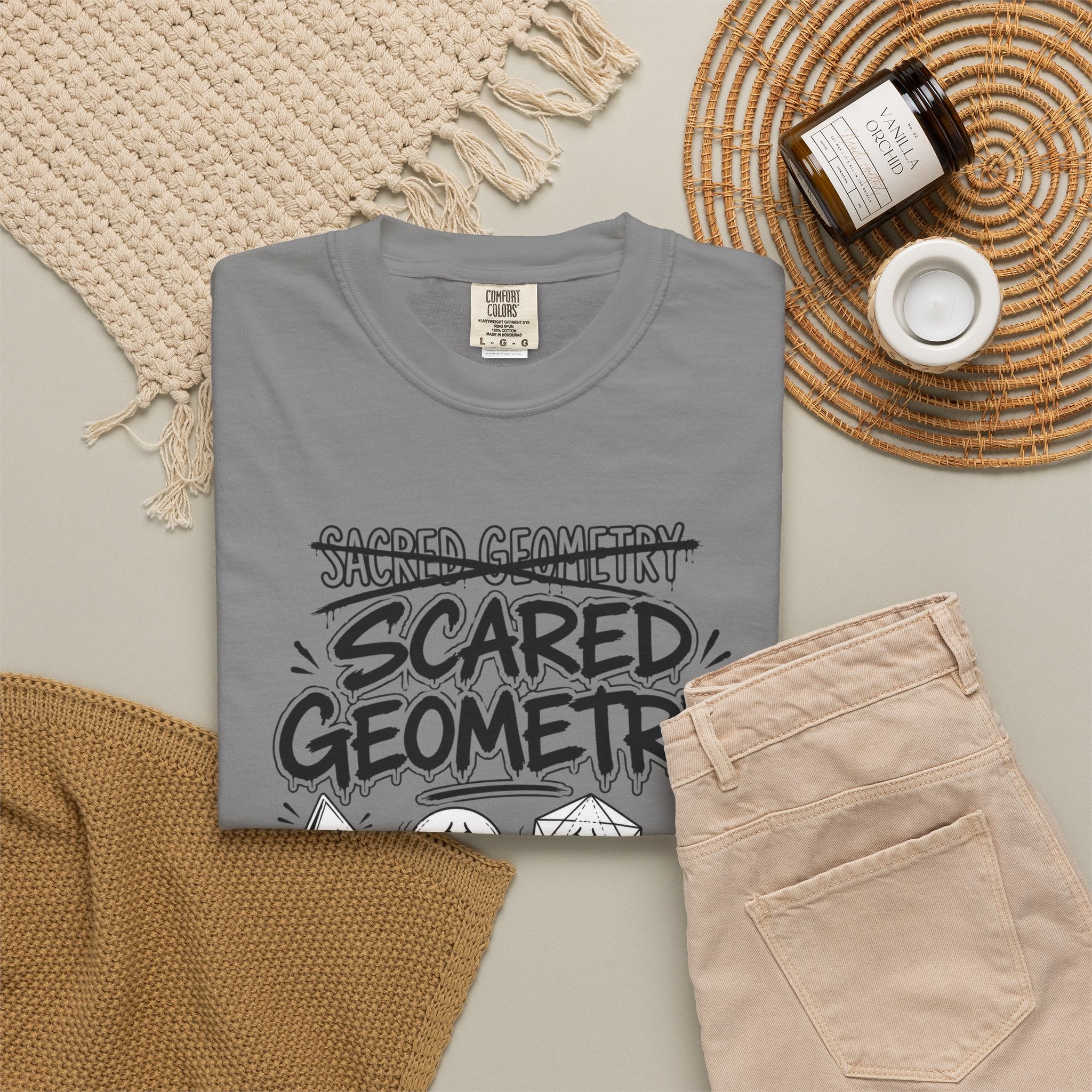 A folded gray 'Scared Geometry' graphic t-shirt with spooky-faced dice designs, styled flat on a knit blanket next to a pair of tan pants.