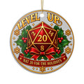 Round hanging ornament with a silver-gray textured background and a gold hanging string. The ornament features a red 20-sided die centered over green holly leaves and red berries, with the words “LEVEL UP” at the top and “NAT 20 FOR THE HOLIDAYS” on a red ribbon at the bottom.