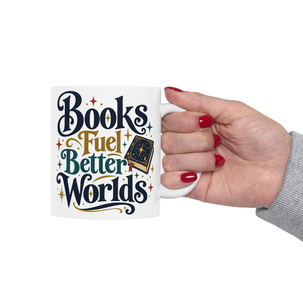 A white ceramic mug with the phrase 'Books Fuel Better Worlds' in stylized navy, gold, and teal lettering, held by a hand with red nail polish against a plain white background.
