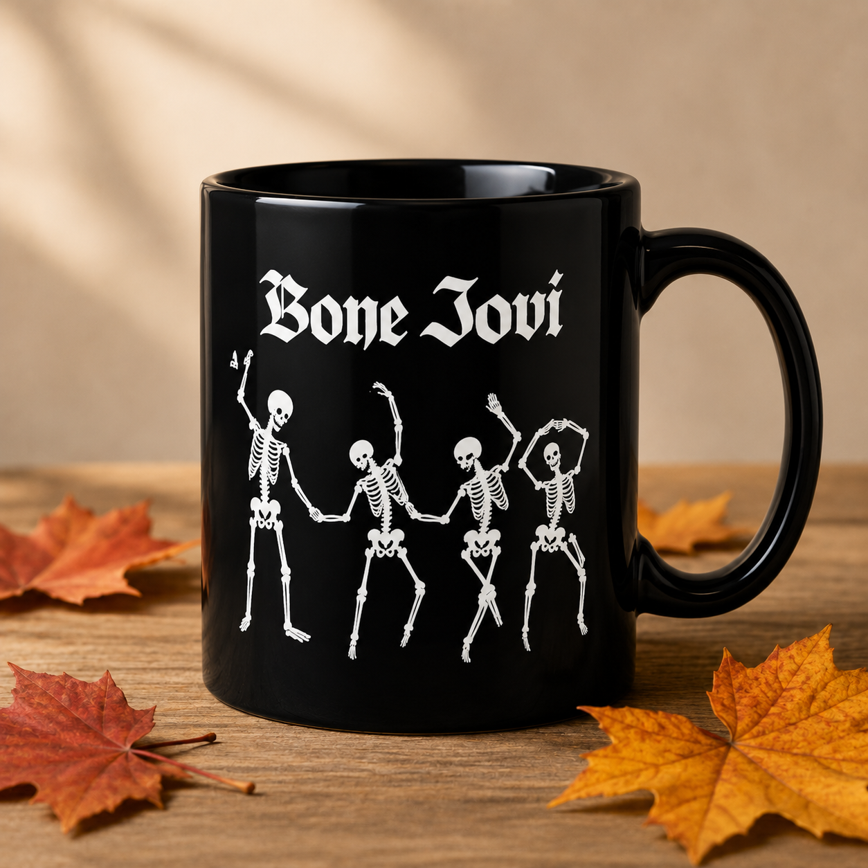 A black ceramic mug with white Gothic-style text reading 'Bone Jovi' above four white dancing skeletons, shown on a wooden surface with autumn leaves and a warm beige background.