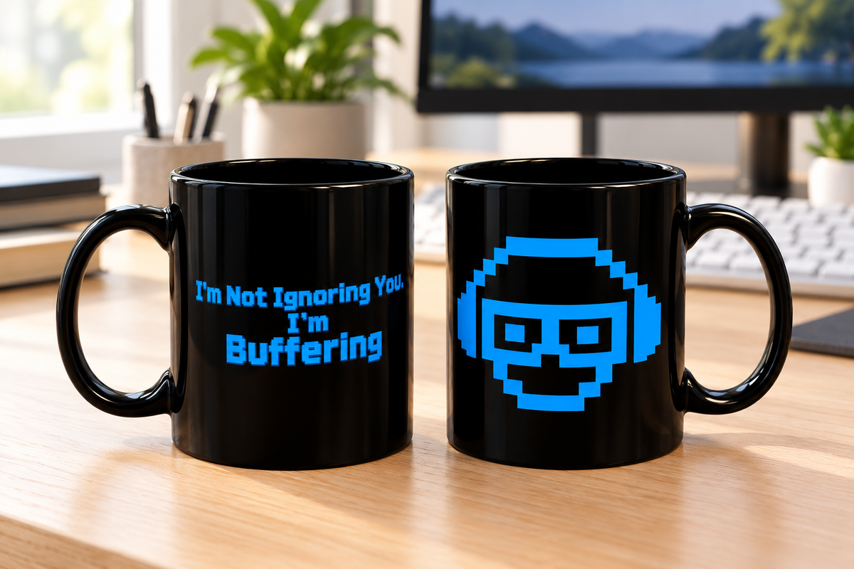 Two black ceramic mugs on a wooden desk in an office setting. The left mug has blue text that reads 'I'm Not Ignoring You, I'm Buffering', and the right mug has a blue pixel-art face graphic.