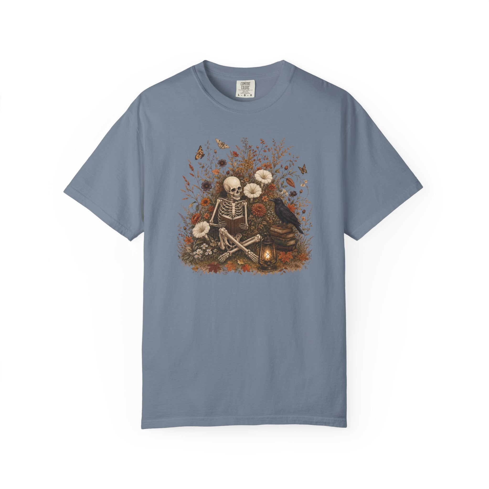 A slate blue short-sleeve t-shirt with a central graphic of a skeleton sitting cross-legged surrounded by white flowers, butterflies, and a crow, set against a plain white background.