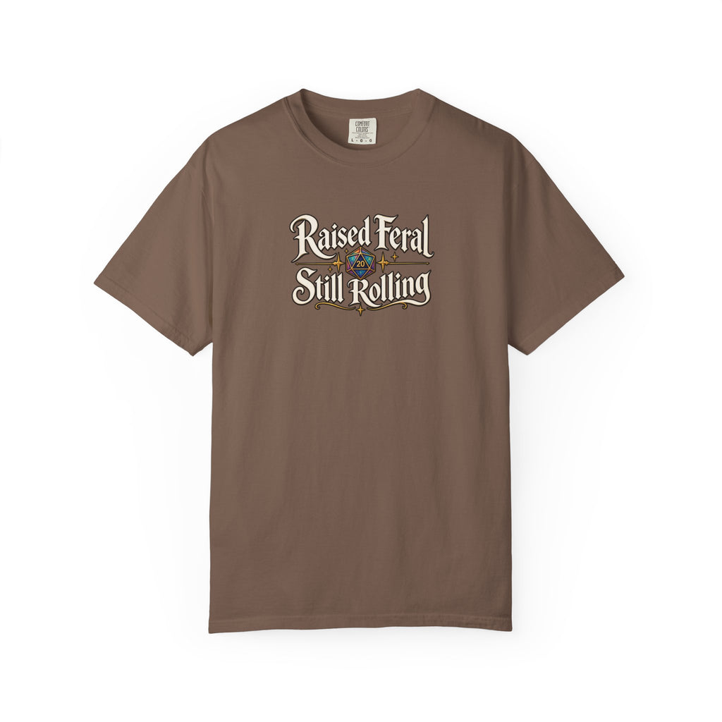 A brown graphic T-shirt  that features the text 'Raised Feral Still Rolling' with a colorful 20-sided die graphic in the center against a white background