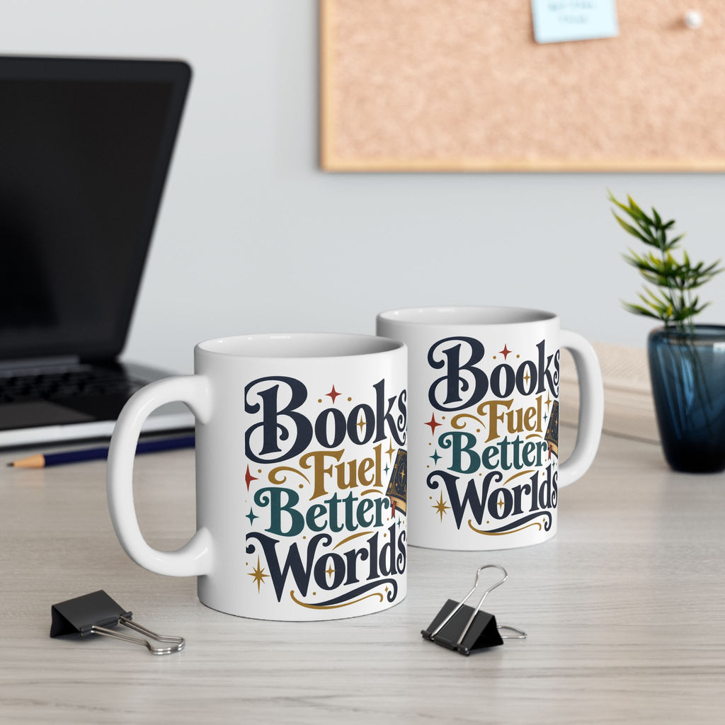 Two white ceramic mugs printed with the phrase 'Books Fuel Better Worlds' in stylized navy, teal, orange, and gold lettering, displayed on a light wood desk with office props including a laptop, binder clips, and a small plant.