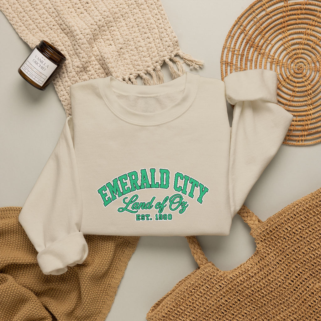 An off-white crewneck sweatshirt with green collegiate-style text reading 'EMERALD CITY Land of Oz EST. 1900', styled in a flat lay with a woven blanket, a candle jar, a woven tray, and a tan tote bag on a light background
