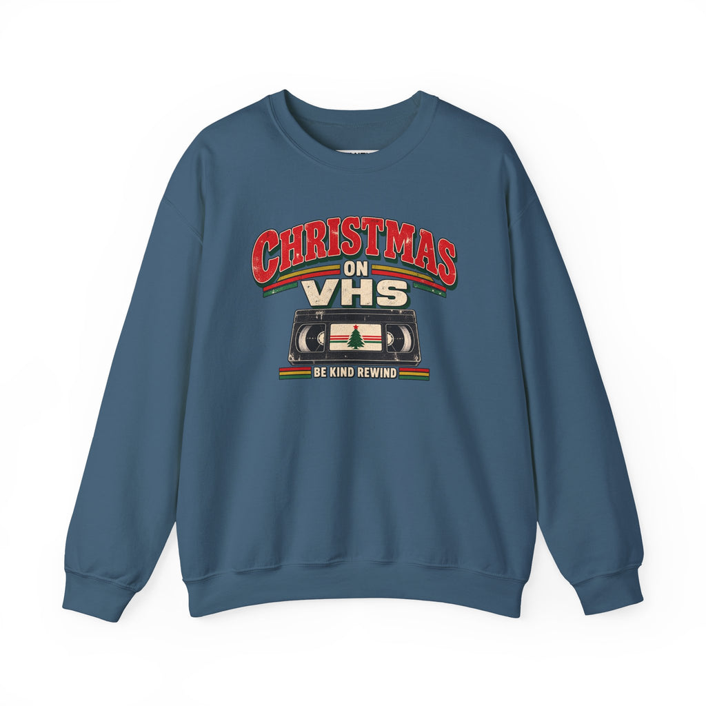 A slate blue crewneck sweatshirt featuring a retro Christmas graphic on the chest that reads 'CHRISTMAS ON VHS' in red and cream lettering above a cassette tape illustration, with 'BE KIND REWIND' underneath.